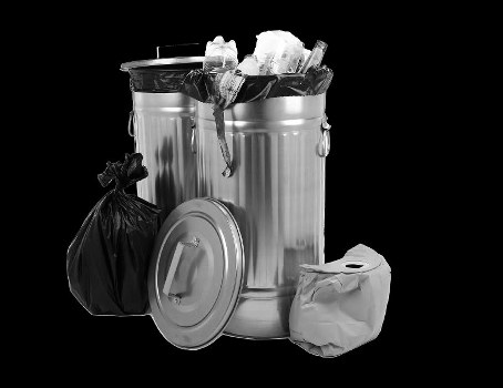 The image shows a large, metallic rubbish bin with a partially open lid, revealing crumpled paper and plastic waste inside. Leaning against the bin is a small black plastic bag, tied at the top, and a round, metallic lid is placed on the ground nearby. To the right of the bin, there is a crumpled white paper bag resting on the paved surface, which appears to be part of a driveway or outdoor area in an urban setting. The background is plain and dark, emphasizing the waste items and the metallic surface of the bin. The scene is consistent with waste collection or rubbish removal activities, typical of services offered by companies like Business Waste Removal Crouch End in the local area, possibly in the N8 postal code district. The overall environment suggests an outdoor space designated for rubbish disposal, with clear focus on refuse contained in the metal bin and associated waste packaging, indicative of domestic or commercial waste management processes.