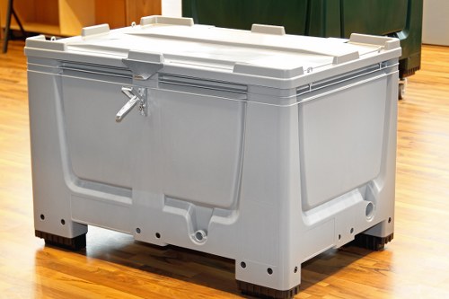 A large, grey plastic wheeled rubbish container with a rectangular shape, situated indoors on a wooden floor. The container has a textured finish and a hinged lid with secure locking mechanisms, one of which is visible on the front left side. It is designed for waste collection and disposal, commonly used in commercial or residential settings for rubbish removal services. The container’s sides feature reinforced panels for durability, and the black wheels at the base allow for easy mobility. The background includes other bins or storage units, indicating an area dedicated to waste management, possibly within a shed or storage room. The scene is well-lit, highlighting the container's sturdy construction and clean surface, emphasizing its role in efficient rubbish removal in the local area.