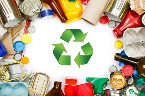 A collection of various waste containers and recyclable materials arranged in a circular pattern around a central green recycling symbol. The items include aluminium cans, plastic bottles, cardboard tubes, and food packaging, with textures ranging from smooth metal surfaces to crinkled paper and plastic. The colours of the waste items are diverse, featuring metallic silver, vibrant reds, blues, yellows, greens, and neutral tones of brown and beige. The background is plain white, emphasizing the waste materials and recycling icon, suggesting emphasis on waste segregation and recycling services. The scene highlights the importance of proper rubbish disposal and recycling practices, relevant to local waste management efforts in the Crouch End area, where Business Waste Removal Crouch End operates, focusing on environmentally responsible rubbish collection and waste disposal solutions.
