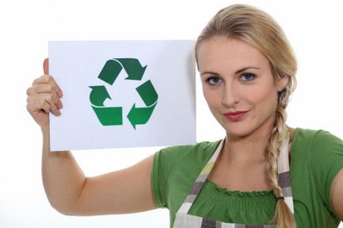 A young woman with long blonde hair styled in a side braid, wearing a green T-shirt and a grey and white apron, holds up a white rectangular sign featuring a green recycling symbol composed of three arrows forming a loop. She is standing outdoors against a plain white background, and her facial expression is neutral and composed. This image relates to waste management and recycling initiatives, emphasizing environmentally responsible rubbish disposal, which aligns with the services offered by Business Waste Removal Crouch End in North London. The woman's posture and the clear, well-lit image focus on the recycling symbol, promoting eco-friendly rubbish removal practices in the local area.