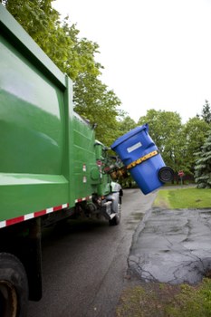 A large green rubbish collection truck parked on a residential street with a blue wheelie bin being lifted and emptied into the vehicle's rear compartment. The truck's textured, painted metal surface reflects natural daylight, and the bin is positioned at an angle, showing its blue plastic body and black wheels. The street features a paved driveway and a grassy verge with trees in the background, indicating an outdoor environment typical of a suburban area. The scene is focused on waste collection, consistent with rubbish removal services provided by Business Waste Removal Crouch End in North London, with the truck partially blocking the view of the street ahead. Overcast sky provides diffuse lighting, highlighting the textures of the bin, the truck's metal surface, and the surrounding greenery.